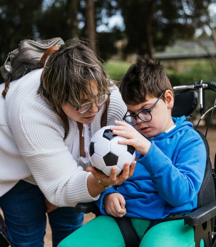 Child with multiple disabilities in a wheelchair playing with a soccer ball with her mother outdoors. people and childhood concept.