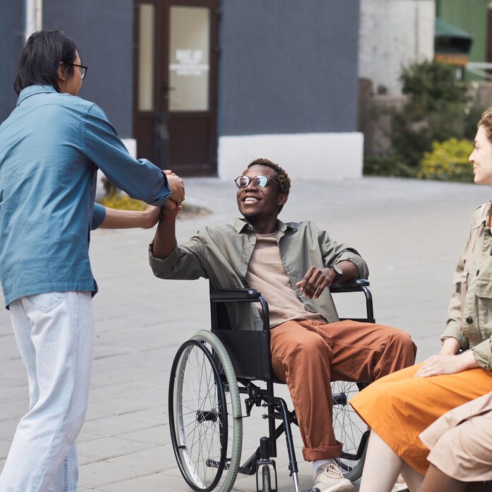 Group of young multi-ethnic friends including disabled man in wheelchair gathering together outdoors, men making handshake