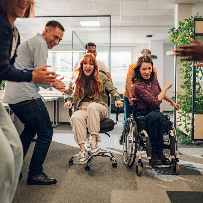 Diverse business team racing on office chairs in an office. Portrait of a redhead female and a woman with disability having fun and racing each other. Multiracial team cheering for both of them.