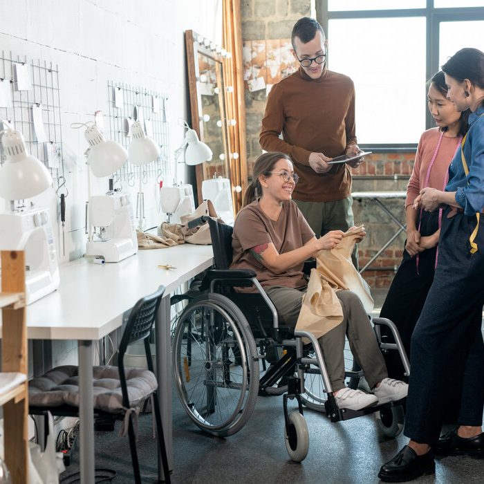 Group of young fashion designers surrounding disable seamtsress in wheelchair during discussion of the main points of new collection