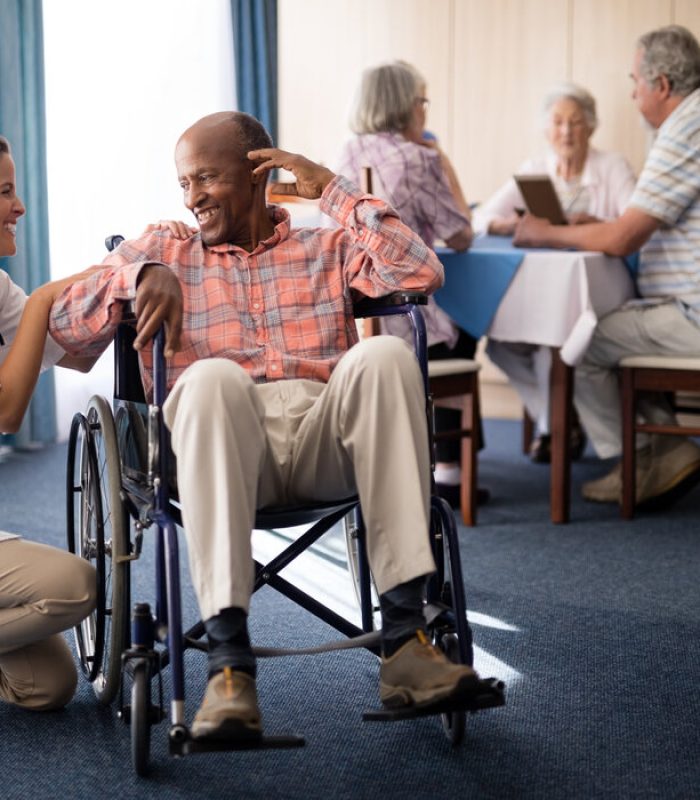 Smiling female doctor kneeling by disabled senior man sitting on wheelchair at retirement home