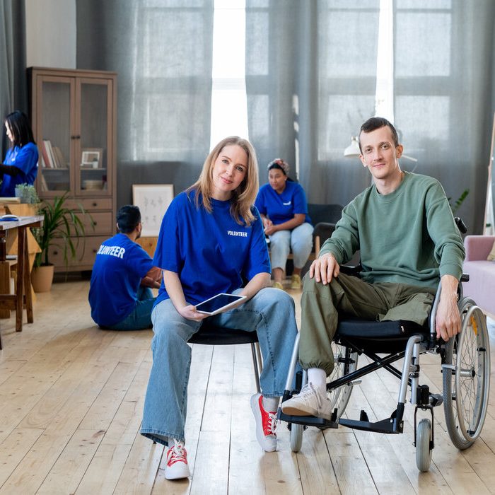 Young blond female sitting by disabled guy in wheelchair against group of volunteers